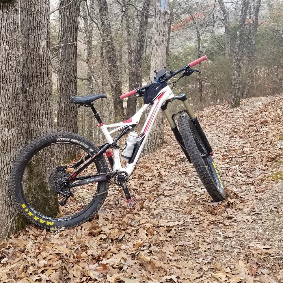 A mountain bike leaning against a tree on a leaf-covered trail in a wooded area. The bike features a white frame with red and black accents and is equipped with a water bottle. The trail is surrounded by tall trees and fallen leaves, suggesting a natural outdoor setting. Hobbs State Park Conservation Area mountain bike trail.