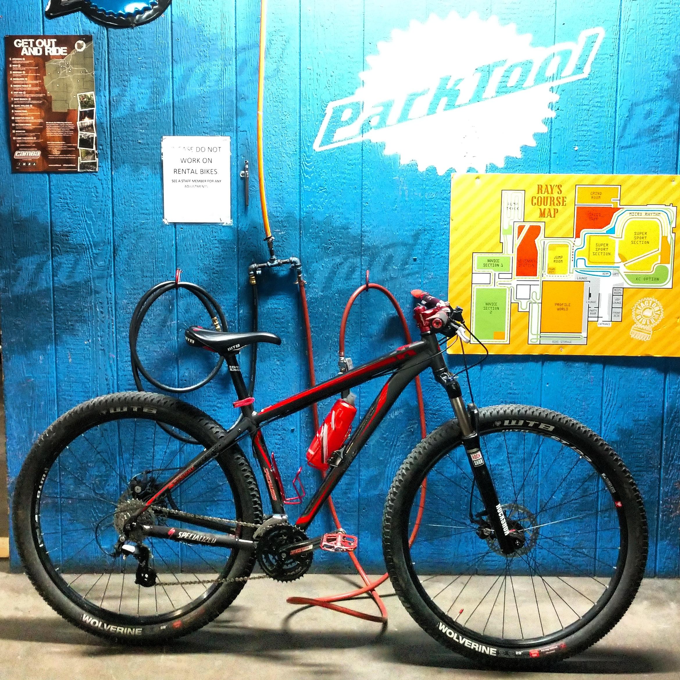 A mountain bike leaning against a blue wall, with a Park Tool logo and a map of Ray's Course displayed nearby. The bike features wide tires, a water bottle holder, and is equipped for trail riding. Additional tools and equipment can be seen in the background. Rays Indoor Mtb Park mountain bike trail.
