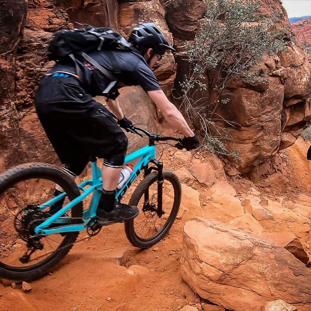 A mountain biker navigating rocky terrain, leaning forward while riding a bright blue bike with thick tires. The surrounding landscape features red rock formations and sparse vegetation, showcasing an adventurous outdoor environment. Hiline mountain bike trail.