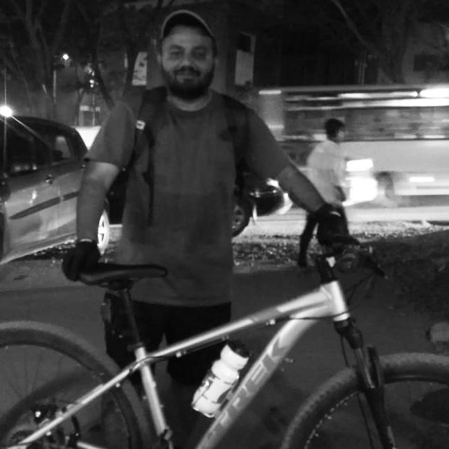 Trek Trek Marlin 4 (29er): A man stands beside a mountain bike in a dimly lit street. He is wearing a gray t-shirt, gloves, and a cap, and has a backpack slung over one shoulder. The background features blurred cars and pedestrians, suggesting an urban environment at night. The image is in black and white.