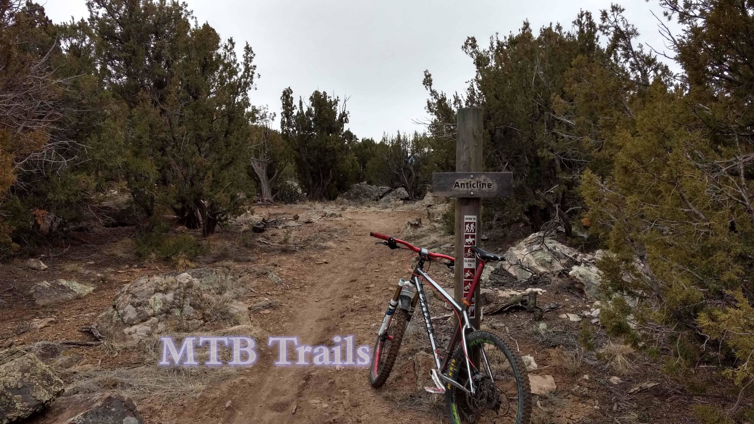 Mountain bike leaning against a trail sign labeled "Anticline," surrounded by rocky terrain and scattered greenery. The path is dirt and leads into a wooded area, indicating a mountain biking trail. The image includes the text "MTB Trails." Oil Well Flats mountain bike trail.
