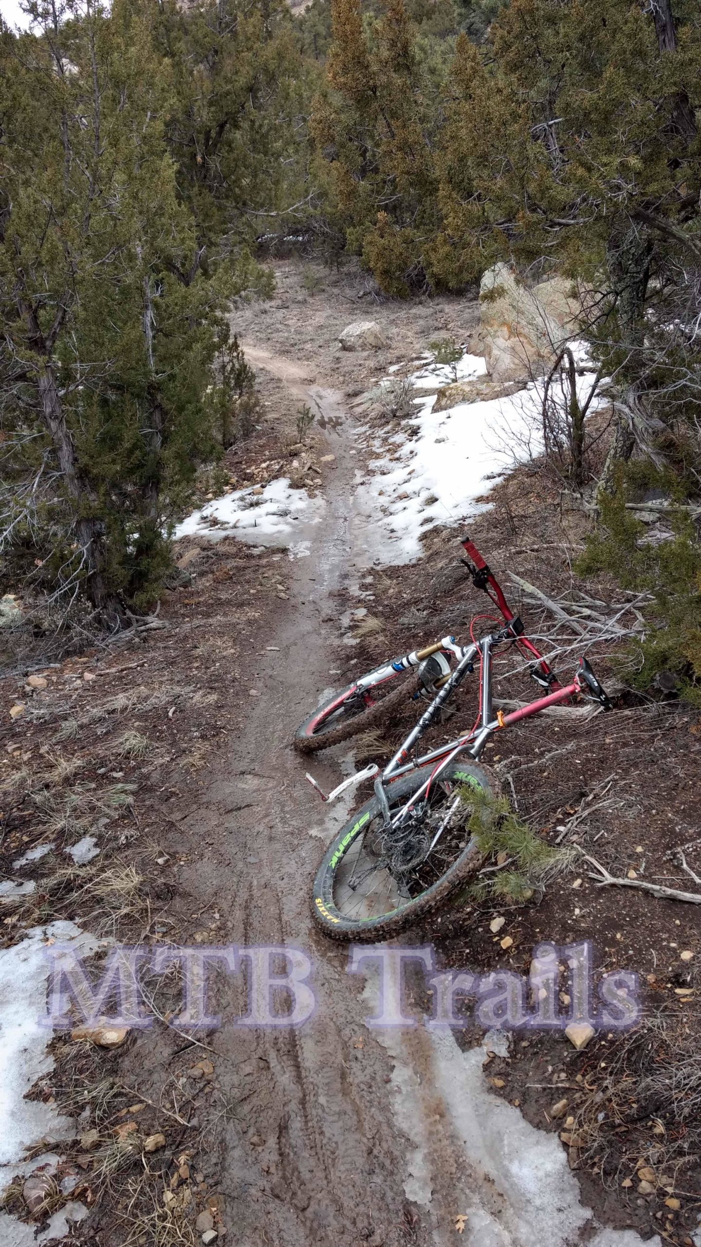 A mountain bike lies on its side on a muddy trail surrounded by trees, with patches of snow visible on the ground. The path winds through a natural landscape, indicating a popular area for mountain biking. Oil Well Flats mountain bike trail.