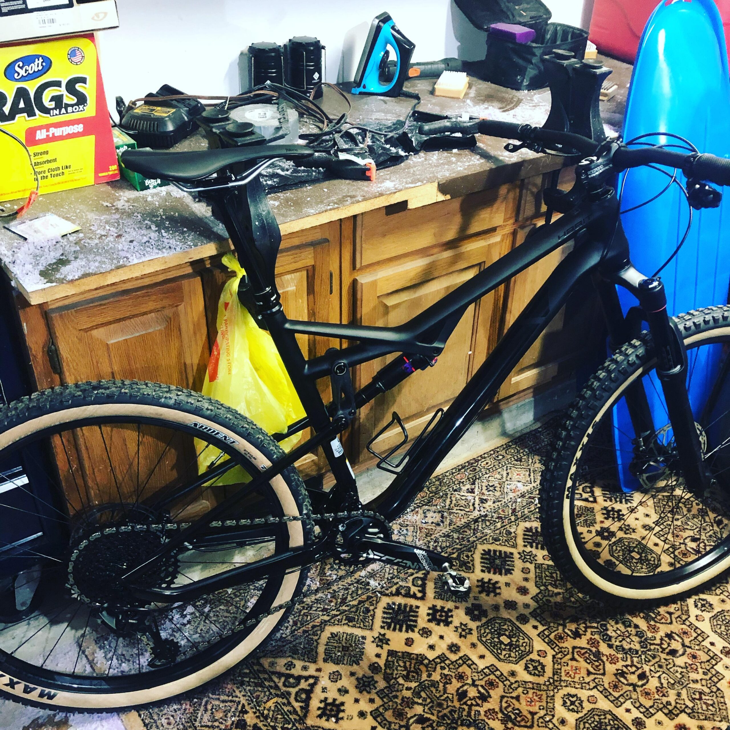 Cannondale Habit Carbon SE: A black mountain bike with tan tires is leaning against a wooden workbench cluttered with tools and equipment. In the background, there is a yellow bag and a blue surfboard. The floor features a patterned rug, and the scene appears to be a garage or workshop setting.