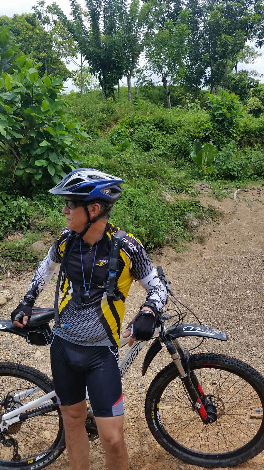 Santa Cruz Superlight: A cyclist wearing a helmet and athletic gear stands next to a mountain bike, surrounded by lush greenery. The cyclist is holding the bike and looking to the side, with gravel and an unpaved path visible in the foreground.