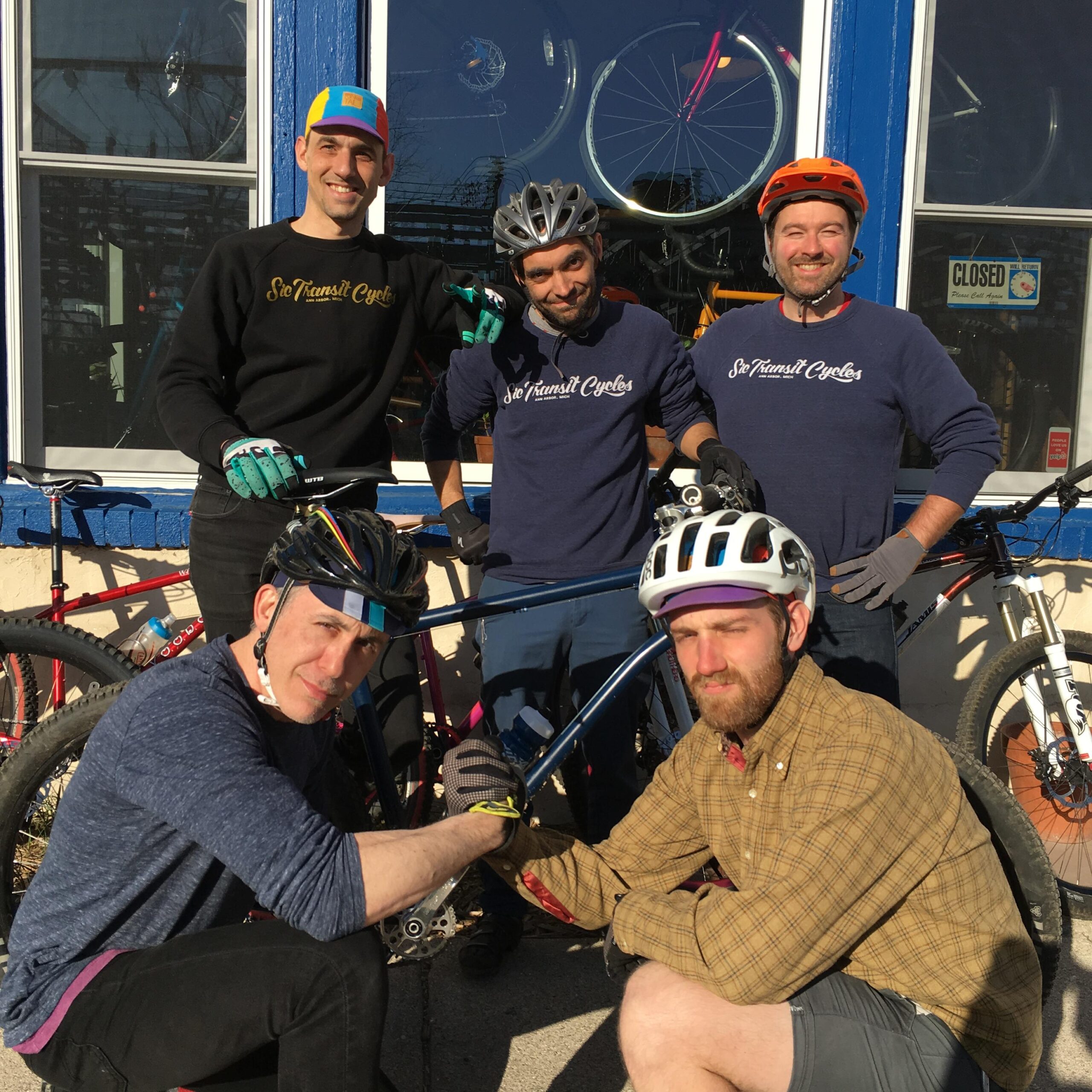 A group of five male cyclists poses outside a bicycle shop, showcasing their bikes. They are dressed in cycling attire, including helmets, gloves, and casual clothing. The shop features colorful bicycle wheels displayed in the background, and the sign on the door indicates it is closed. The men appear to be smiling and enjoying each other