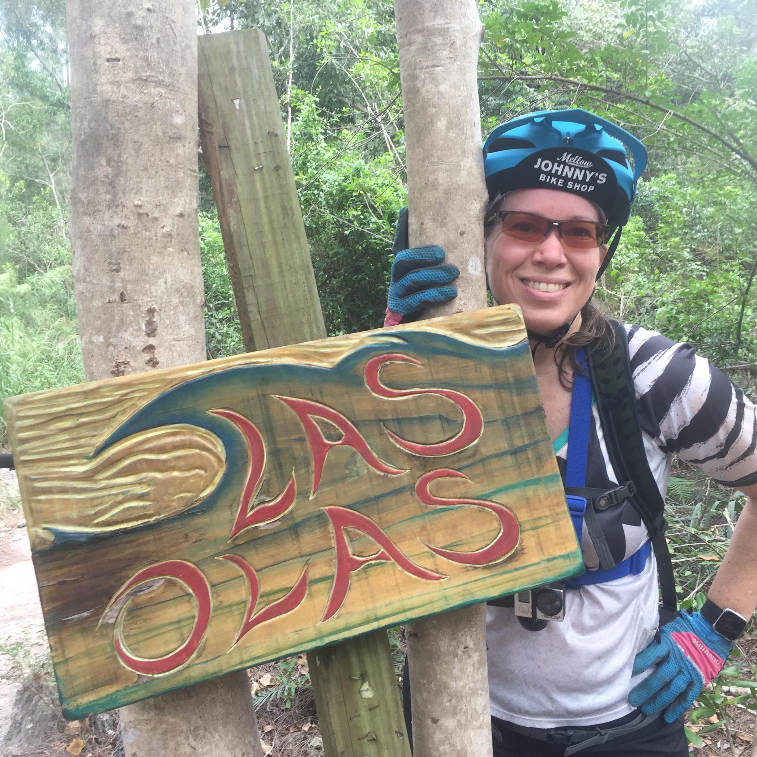 A person wearing a helmet and colorful gloves stands next to a wooden sign that reads "Las Olas," surrounded by trees and greenery. The individual is smiling, and their bike gear is visible. Markham Park mountain bike trail.
