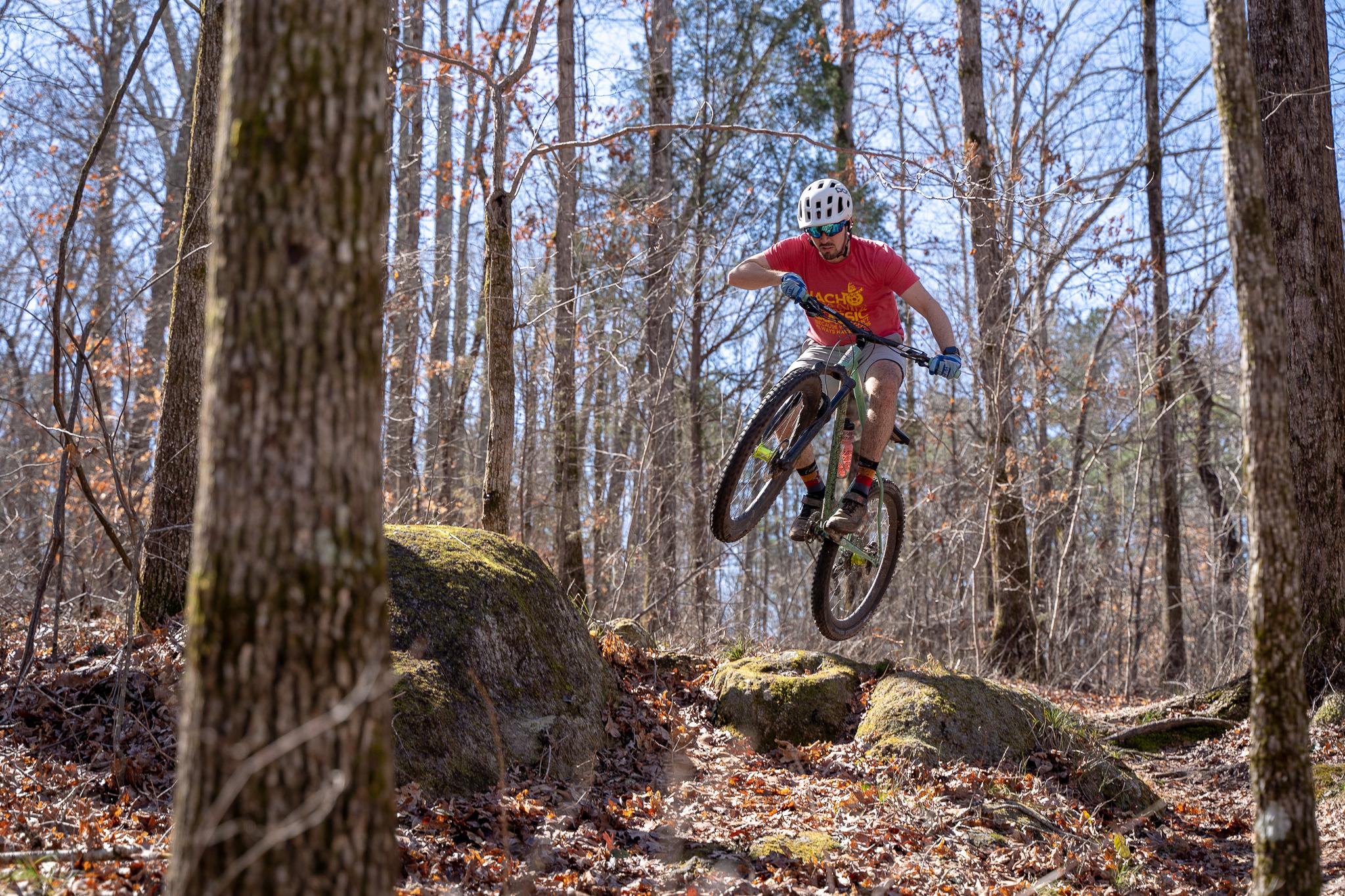 A mountain biker performing a jump over a rocky terrain, surrounded by trees and fallen leaves in a wooded area. The cyclist is wearing a helmet, gloves, and a colorful t-shirt, capturing an action-packed moment in outdoor cycling. Clinton Nature Preserve mountain bike trail.