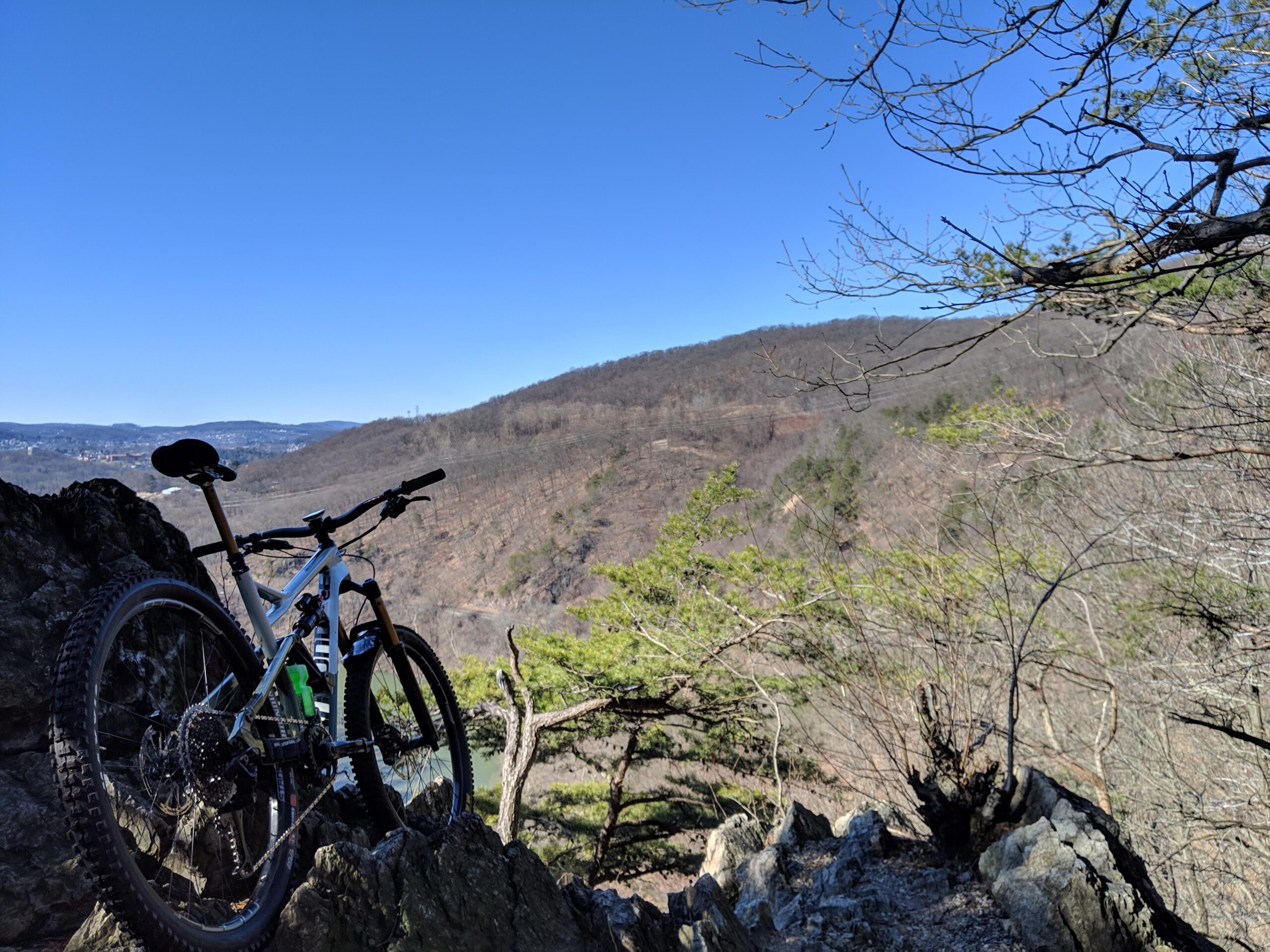 Cannondale Jekyll: A mountain bike resting on a rocky outcrop with a clear blue sky and distant hills in the background. Sparse trees and an expansive landscape are visible, revealing a serene outdoor setting.