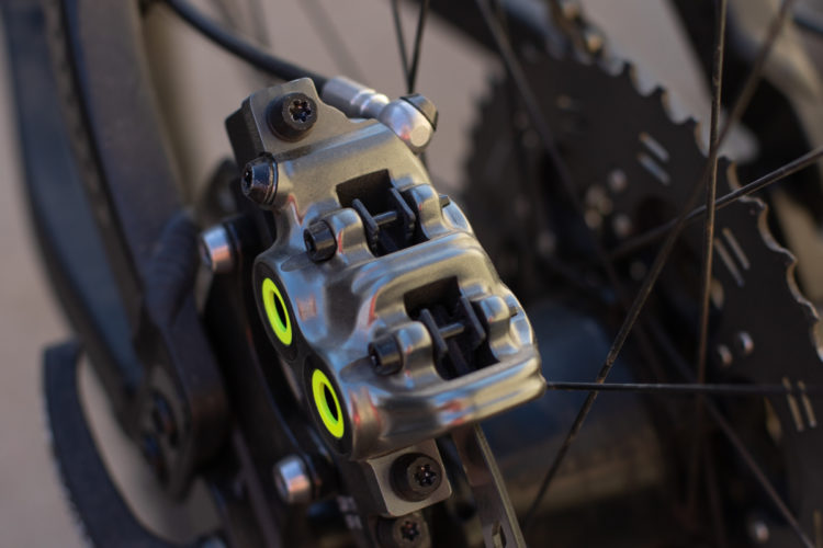 Close-up image of a bicycle brake caliper, showcasing its metallic structure with bright yellow accents, along with surrounding components of the bicycle's wheel and brake system.