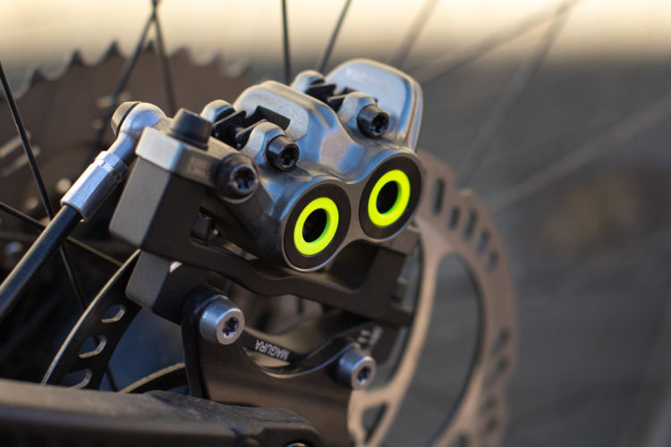 Close-up image of a bicycle brake caliper with bright yellow accents, mounted on a disc brake. The intricate mechanical components and screws are visible, showcasing the precision engineering of the brake system. The background features part of the bicycle wheel and disc rotor, blending into a blurred landscape.
