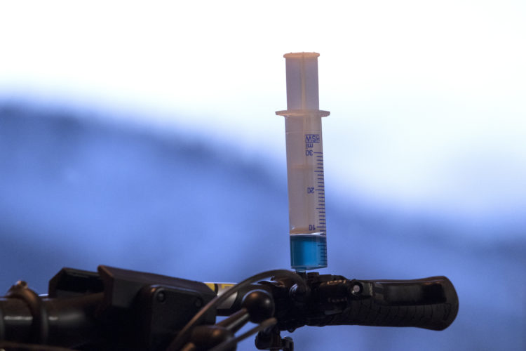 A close-up image of a syringe filled with a blue liquid, positioned upright on the handlebar of a bicycle. The background features a blurred, light-colored gradient.