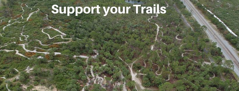 Aerial view of a lush green landscape featuring winding dirt trails surrounded by trees, with a road visible on the right side. The text "Support your Trails" is prominently displayed across the top of the image. Jonathan Dickinson State Park mountain bike trail.