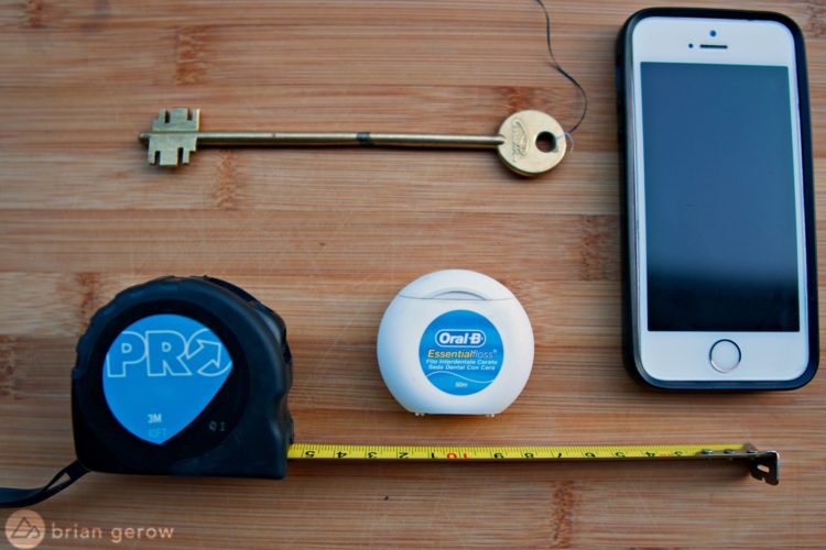 A flat lay image featuring a brass key, a measuring tape with a black casing, a container of Oral-B dental floss, and a smartphone placed on a wooden surface. The measuring tape is partially extended, showing measurements.