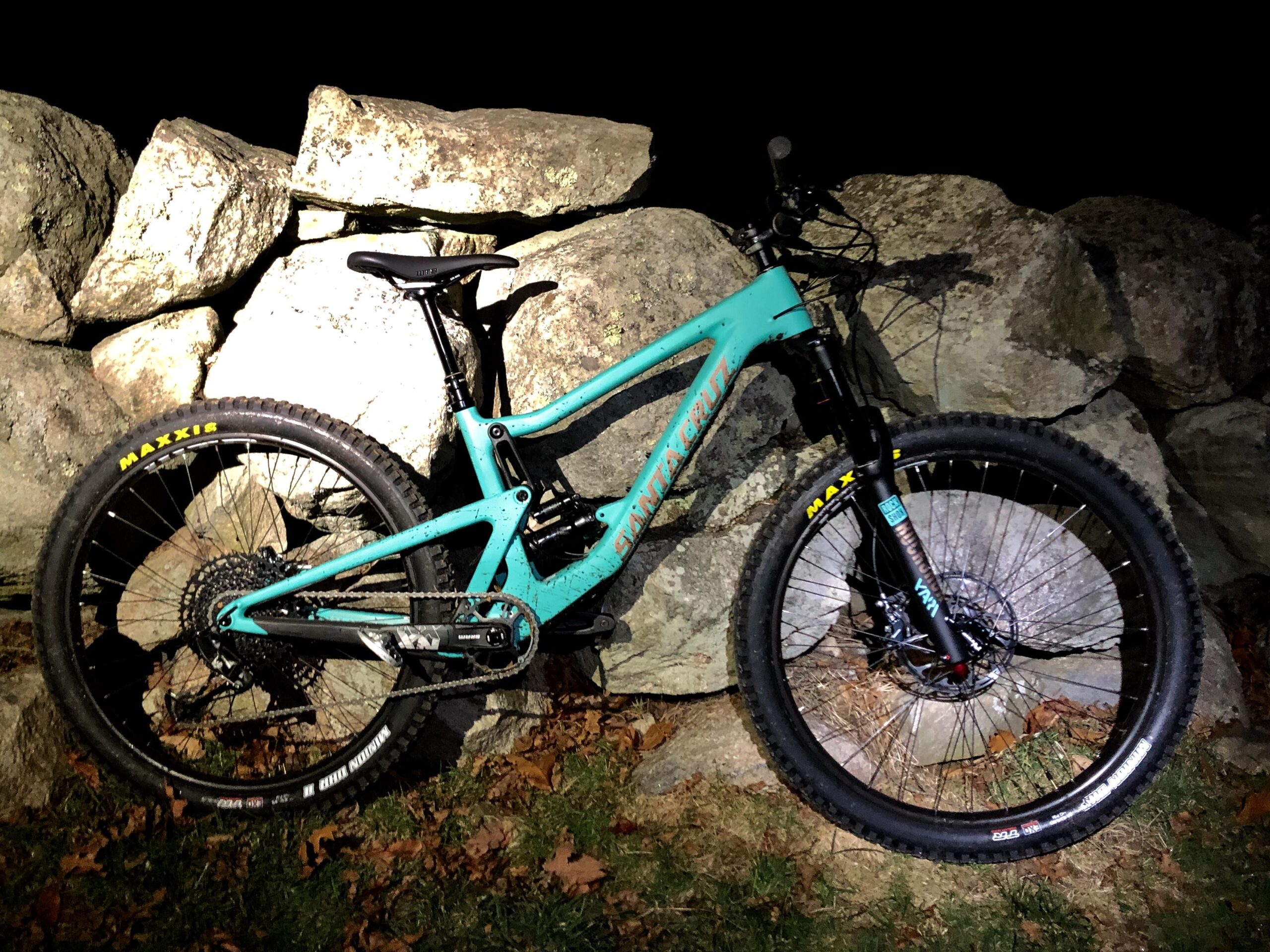Santa Cruz Bronson C: A turquoise mountain bike leaning against a rock wall at night, surrounded by fallen leaves. The bike features large tires with visible branding and has a sleek design suitable for trail riding. The scene is illuminated by a light source, casting shadows on the rocks behind.