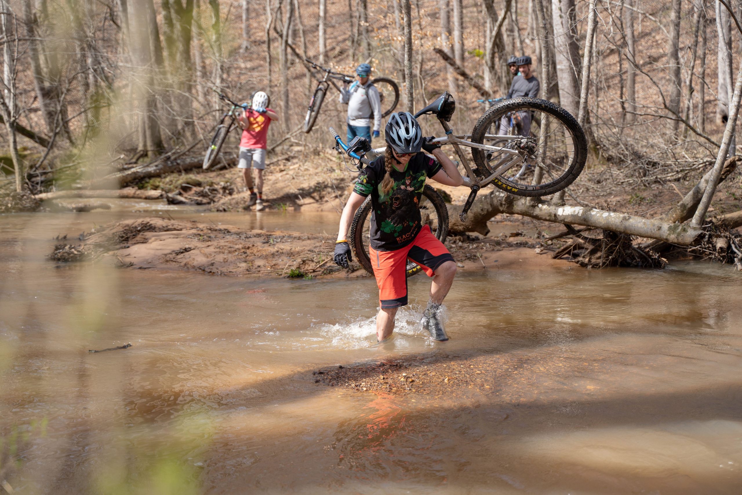 A group of mountain bikers navigating through a shallow stream in a forested area. One rider, dressed in a colorful shirt and shorts, is wading through the water while carrying their bike, while others are visible in the background holding their bikes. The scene captures a moment of adventure and camaraderie in nature. Clinton Nature Preserve mountain bike trail.