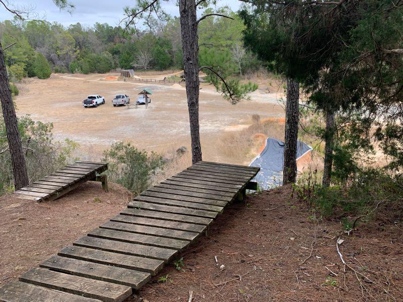 A wooden pathway leading down from a hill, surrounded by pine trees. In the background, an open area with a few parked vehicles and a shelter is visible, along with patches of dry grass and some remnants of landscaping features. The scene is set in a natural, outdoor environment. Santos mountain bike trail.