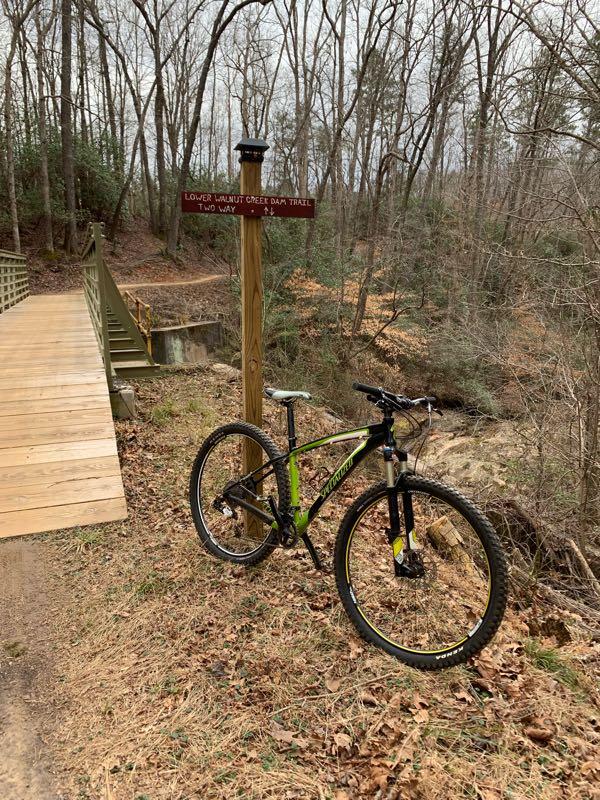 A mountain bike leaning against a wooden sign that reads "Lower Walnut Creek Data Trail" with an arrow indicating a two-way path. The scene is set in a wooded area with bare trees and pathways visible, alongside a wooden bridge leading over a small creek. The ground is covered with fallen leaves. Chicopee Woods mountain bike trail.
