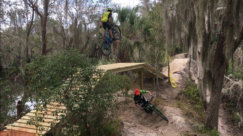 Two mountain bikers navigating a dirt trail in a wooded area. One biker is mid-air, jumping over a wooden ramp, while the second biker is leaning into a corner on the ground. Surrounding vegetation includes trees with Spanish moss and bushes, creating a natural outdoor setting. Loyce E. Harpe Park mountain bike trail.