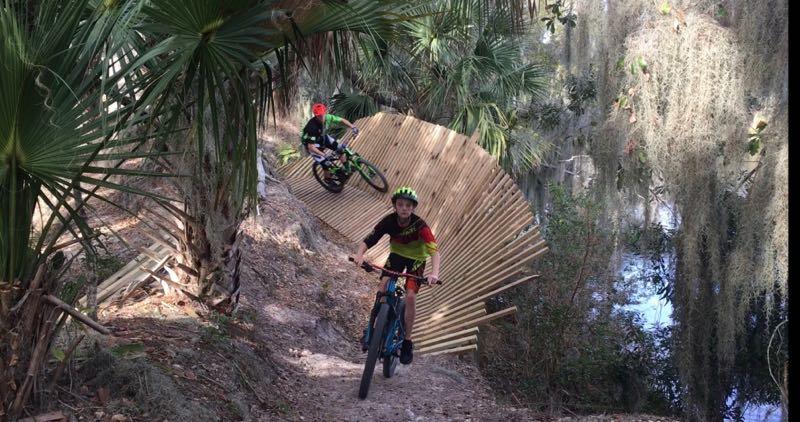 Two young boys riding mountain bikes on a dirt trail surrounded by lush greenery. One boy, wearing a red helmet and green shirt, is seen mid-ride on a wooden ramp, while the other boy in a yellow and red shirt navigates the trail. A peaceful water body reflects sunlight in the background, enhancing the natural setting. Loyce E. Harpe Park mountain bike trail.
