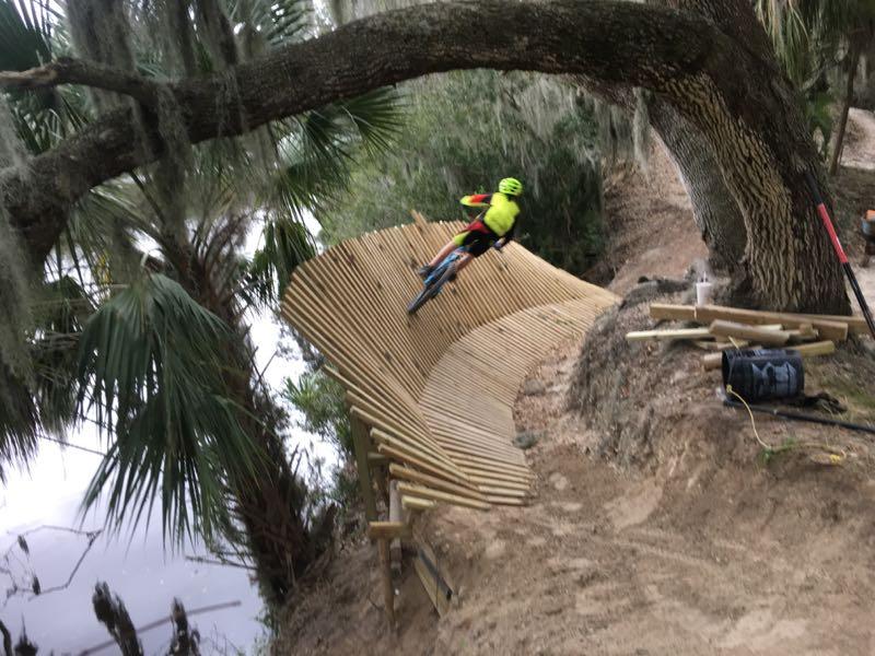 A mountain biker in a bright yellow jacket and helmet rides along a wooden curved ramp, suspended above a sandy bank next to a body of water, surrounded by lush greenery and palm trees. Tools and materials are visible nearby, indicating ongoing construction or trail maintenance. Loyce E. Harpe Park mountain bike trail.