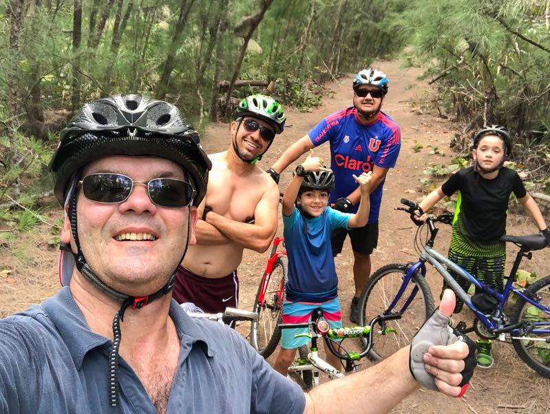 A group of five people, including two adults and three children, take a selfie while mountain biking on a dirt path surrounded by trees. Everyone is wearing helmets, and the adults show smiles and thumbs-up gestures. Two children are sitting on their bikes, while an adult stands behind them, posing with his arms crossed. The scene conveys a sense of fun and outdoor activity. Oleta River State Park mountain bike trail.