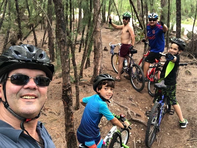 A group of five people, including two children, wearing helmets and smiling, posing for a selfie while on a biking trail surrounded by trees. The adults are on different bicycles, while the children are mounted on their bikes, dressed in casual athletic clothing. The setting is lush with greenery, suggesting an outdoor adventure. Oleta River State Park mountain bike trail.