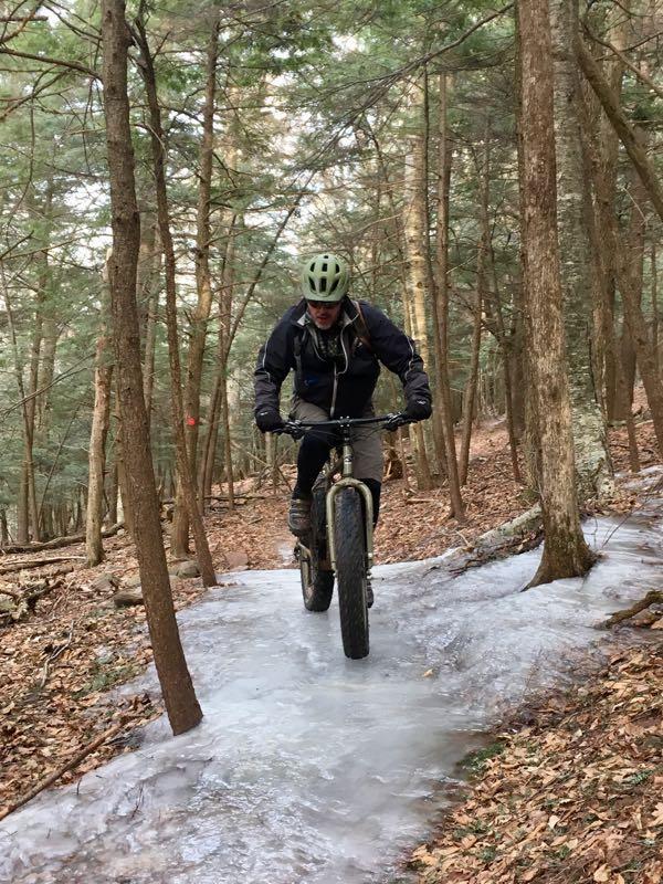A person riding a fat tire bike on a snowy trail through a forest, surrounded by tall trees and scattered leaves on the ground. Elm Ridge mountain bike trail.