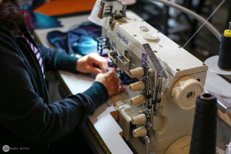 A close-up image of a person sewing at a sewing machine, with hands focused on fabric, surrounded by spools of thread. The setting appears to be an indoor workspace, highlighting the details of the sewing process.