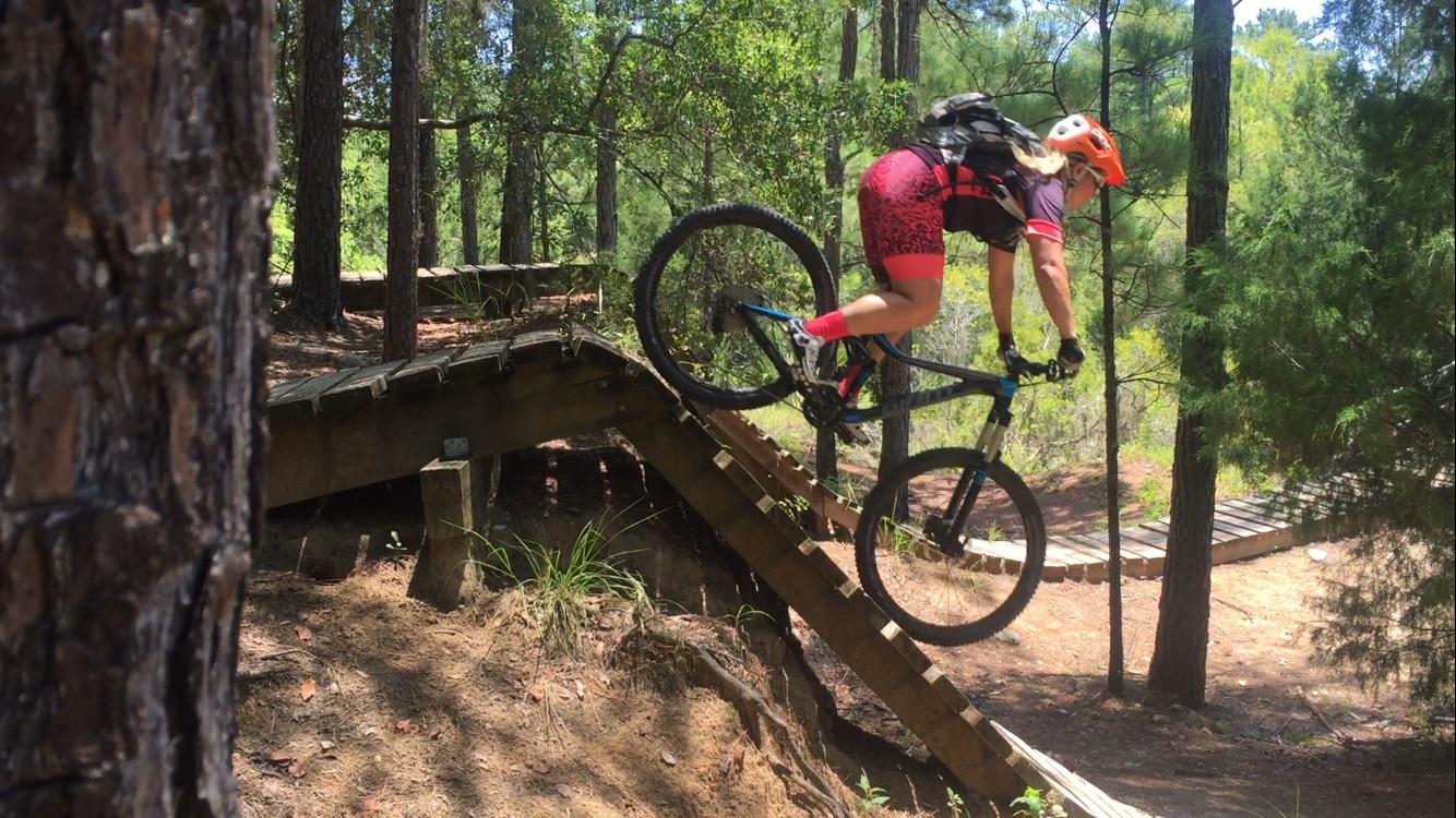 A mountain biker in a bright pink and black outfit descends a wooden ramp while navigating through a forested trail. The biker is airborne, with one wheel raised as they navigate the transition, surrounded by tall trees and natural greenery. Santos mountain bike trail.