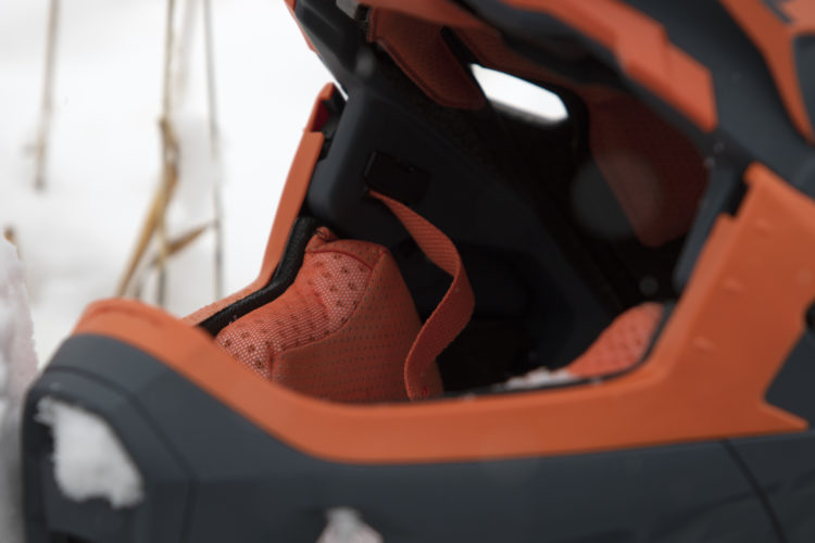 Close-up of the inside of a ski boot, featuring a cushioned orange liner and adjustable straps, set against a snowy background.