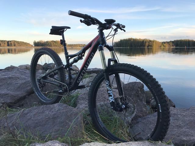 Specialized Stumpjumper FSR 6Fattie: A mountain bike resting on large rocks by a calm lake, surrounded by trees and a clear sky. The sunset casts a warm glow, highlighting the bike's design and the tranquil water.