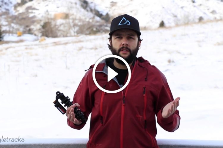 A person in a red jacket stands outdoors in a snowy landscape, holding a bicycle shock absorber in one hand and gesturing with the other. The background features snow-covered hills and trees. A play button overlay indicates that this is a video.