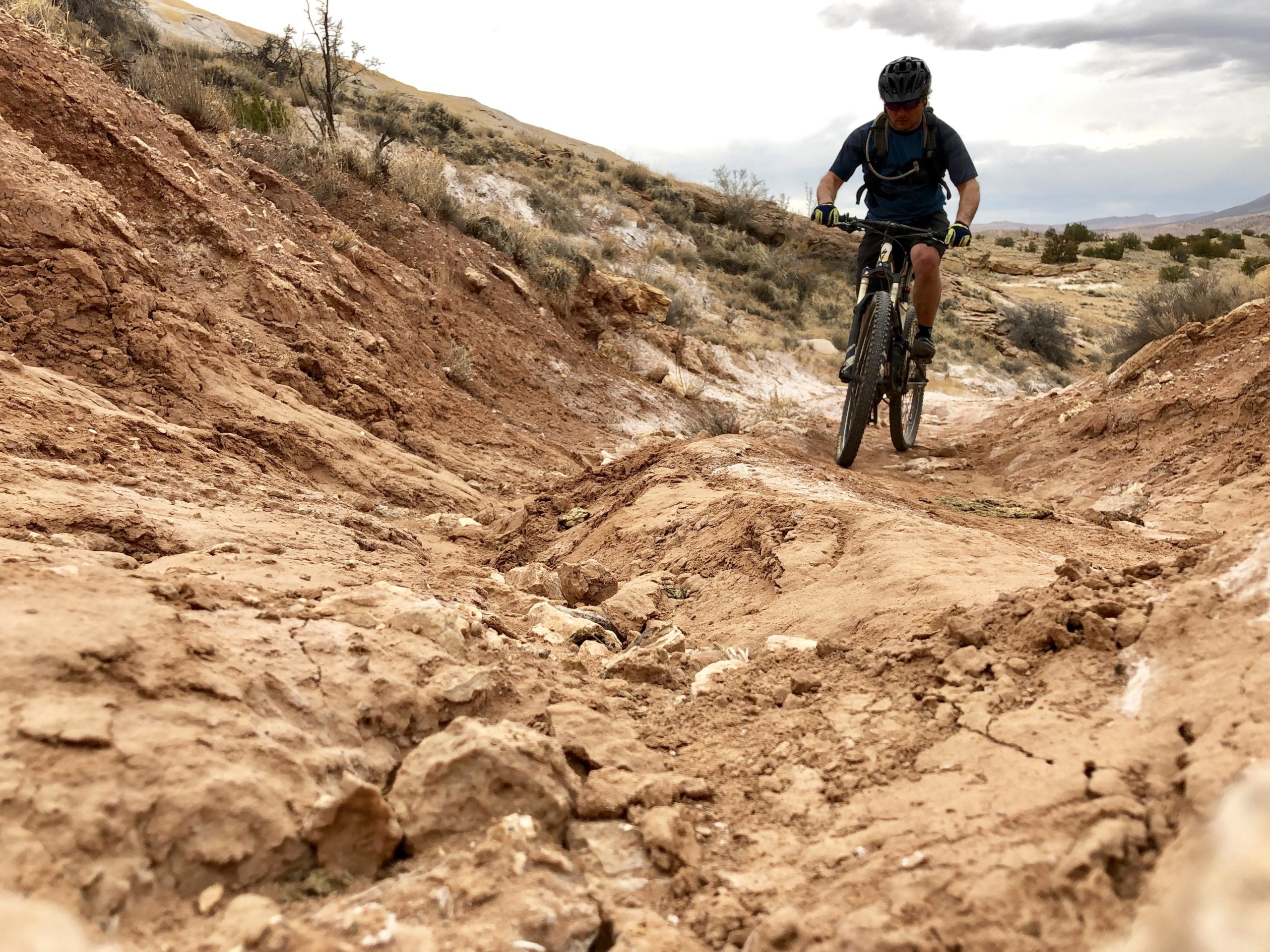 A mountain biker navigating a rugged dirt trail, surrounded by dry vegetation and rocky terrain, with a cloudy sky above. The focus is on the biker, who is wearing a helmet and riding downhill on a mountain bike. White Ridge Bike Trails mountain bike trail.