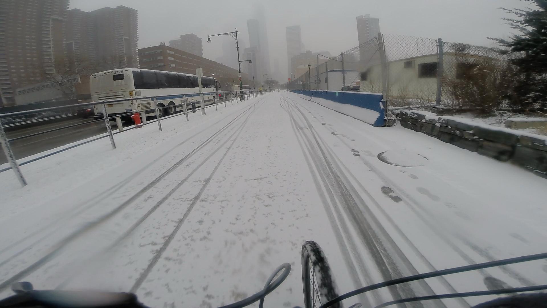 A snowy bike path in an urban area, with tire tracks and footprints in the snow. A bus is visible on the road next to the path, while tall buildings are partially obscured by fog in the background. West Street Greenway mountain bike trail.
