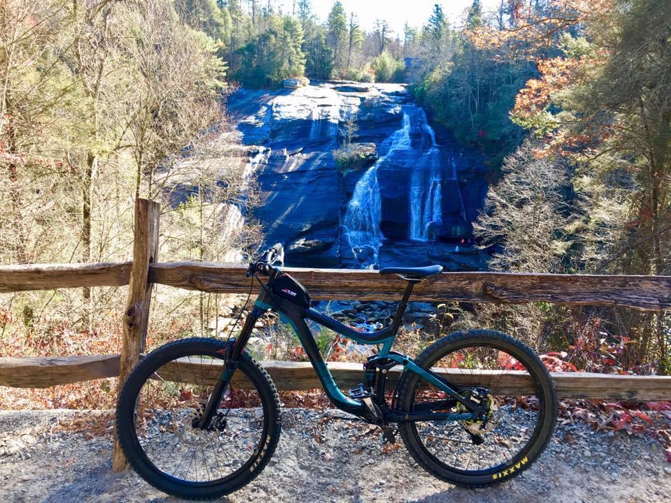 A mountain bike resting against a wooden fence, with a scenic view of a waterfall cascading down rocky terrain, surrounded by trees in autumn foliage. DuPont State Forest mountain bike trail.