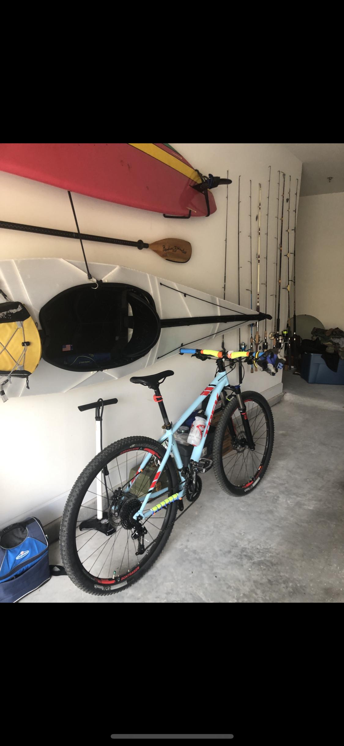 Trek Trek x caliber 8: A garage scene featuring a light blue mountain bike with red and yellow accents standing alongside various outdoor gear. In the background, there are kayaks and a paddle mounted on the wall, as well as multiple fishing rods hanging from hooks. A pump and a cooler bag are also visible on the floor, indicating an active lifestyle focused on biking, kayaking, and fishing.