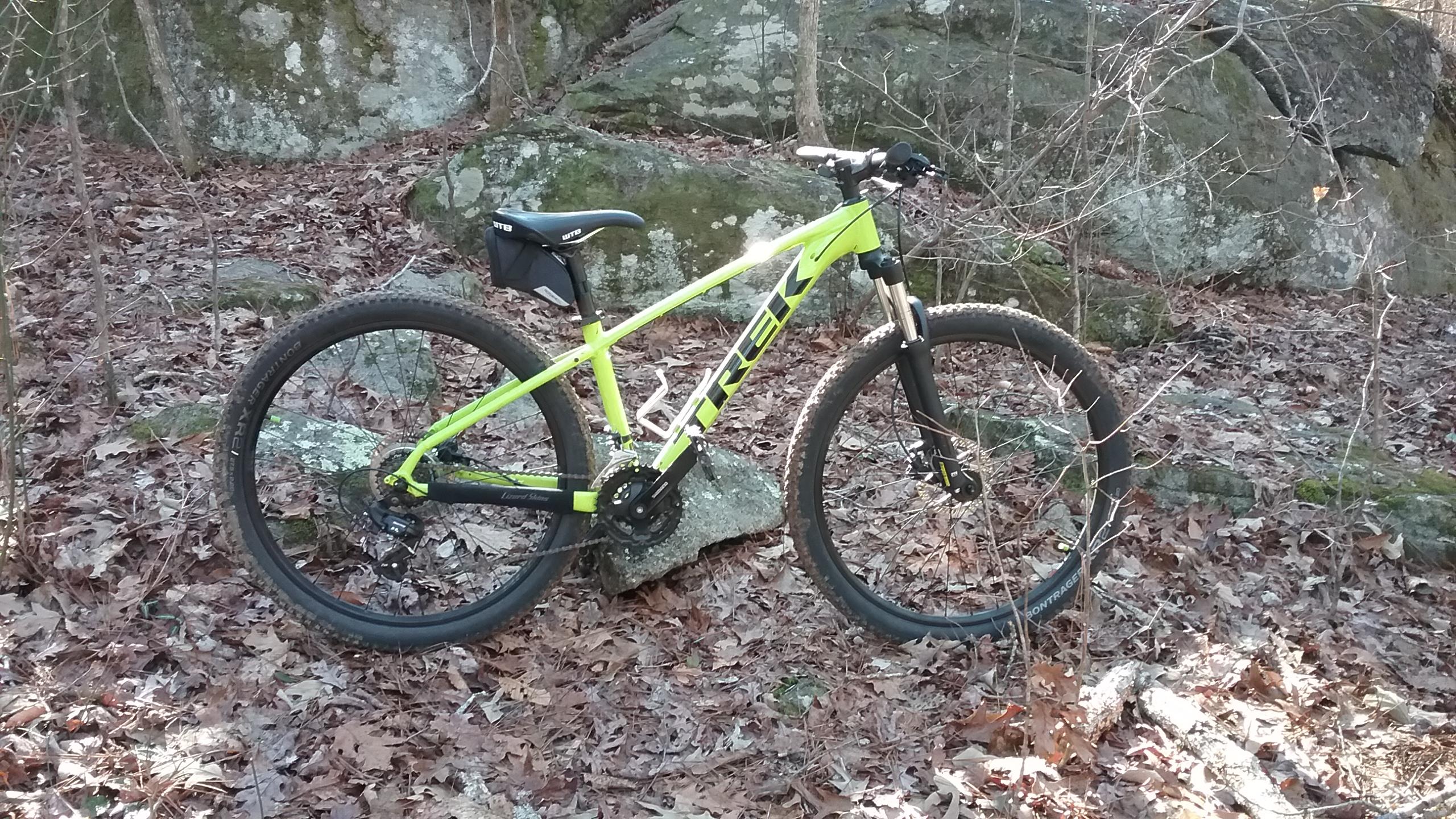 Trek Marlin 5: A bright green mountain bike parked on a rocky surface surrounded by fallen leaves and trees in a wooded area.