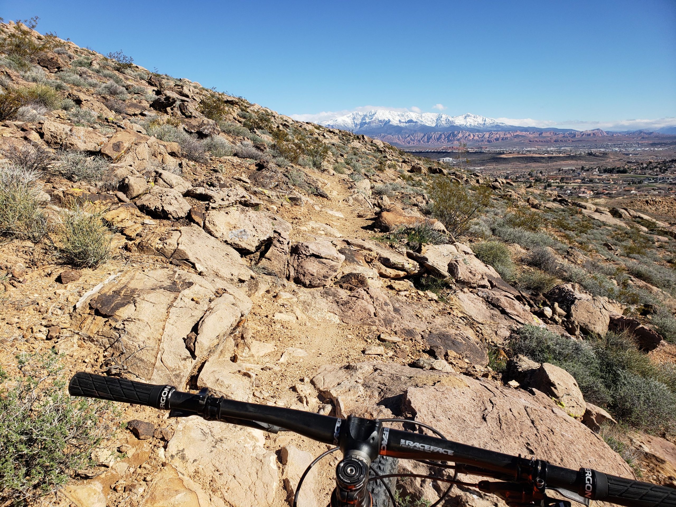 A mountain bike handlebar is shown in the foreground, positioned above a rocky trail. The background features a scenic view of a mountainous landscape with snow-capped peaks, under a clear blue sky. Sparse vegetation covers the rocky terrain, indicating a rugged outdoor environment ideally suited for biking or hiking. Kentucky Lucky Chicken mountain bike trail.