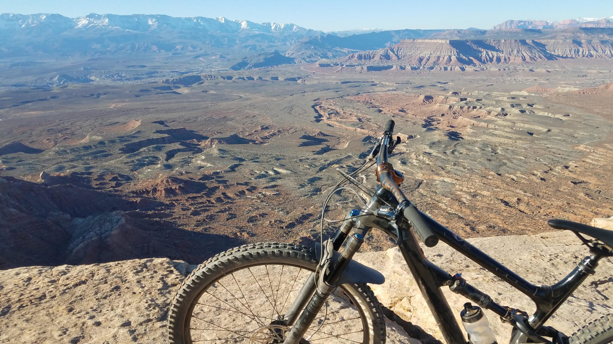 A mountain bike rests on the edge of a rocky overlook, with a vast expanse of rugged terrain and distant snow-capped mountains in the background. The landscape features various geological formations, showcasing a mix of arid earth tones and textures under a clear sky. Gooseberry Mesa mountain bike trail.