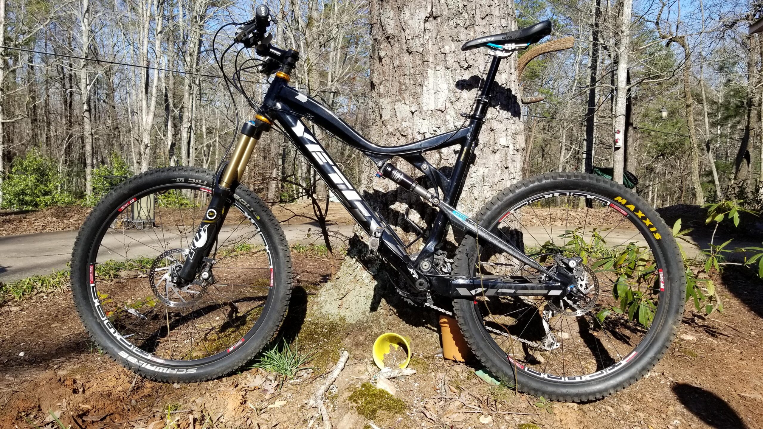 Yeti 575: A black mountain bike is leaning against a tree in a forested area. The bike features a prominent front suspension fork and wide tires. Surrounding the bike are patches of earth, moss, and small plants, with a yellow bowl partially buried in the ground. In the background, a path and additional trees are visible, indicating a natural outdoor setting.