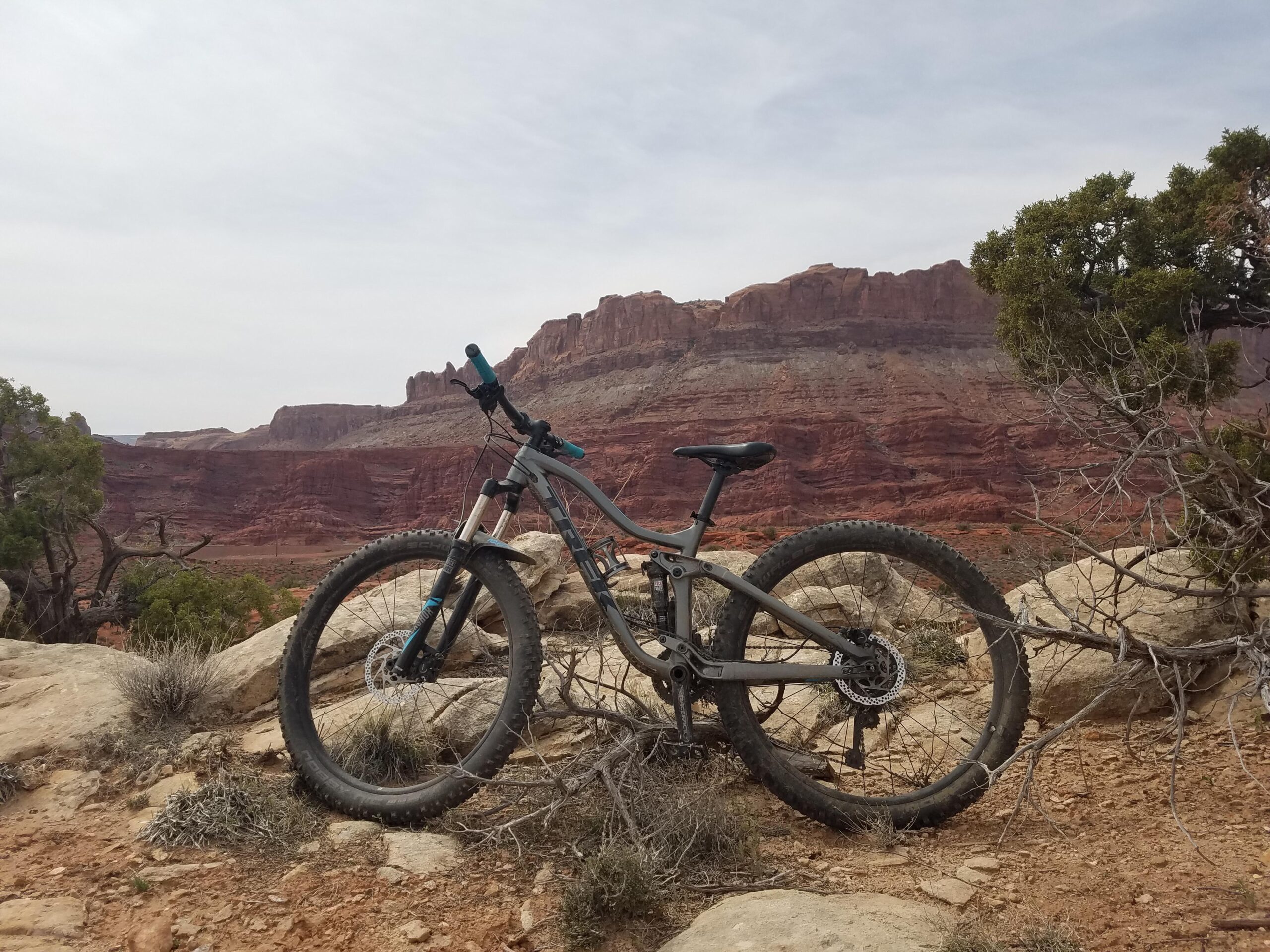 Trek Fuel EX: A mountain bike leaning against rocks with a scenic view of red rock formations in the background. The landscape features dry vegetation and rugged terrain under a cloudy sky.