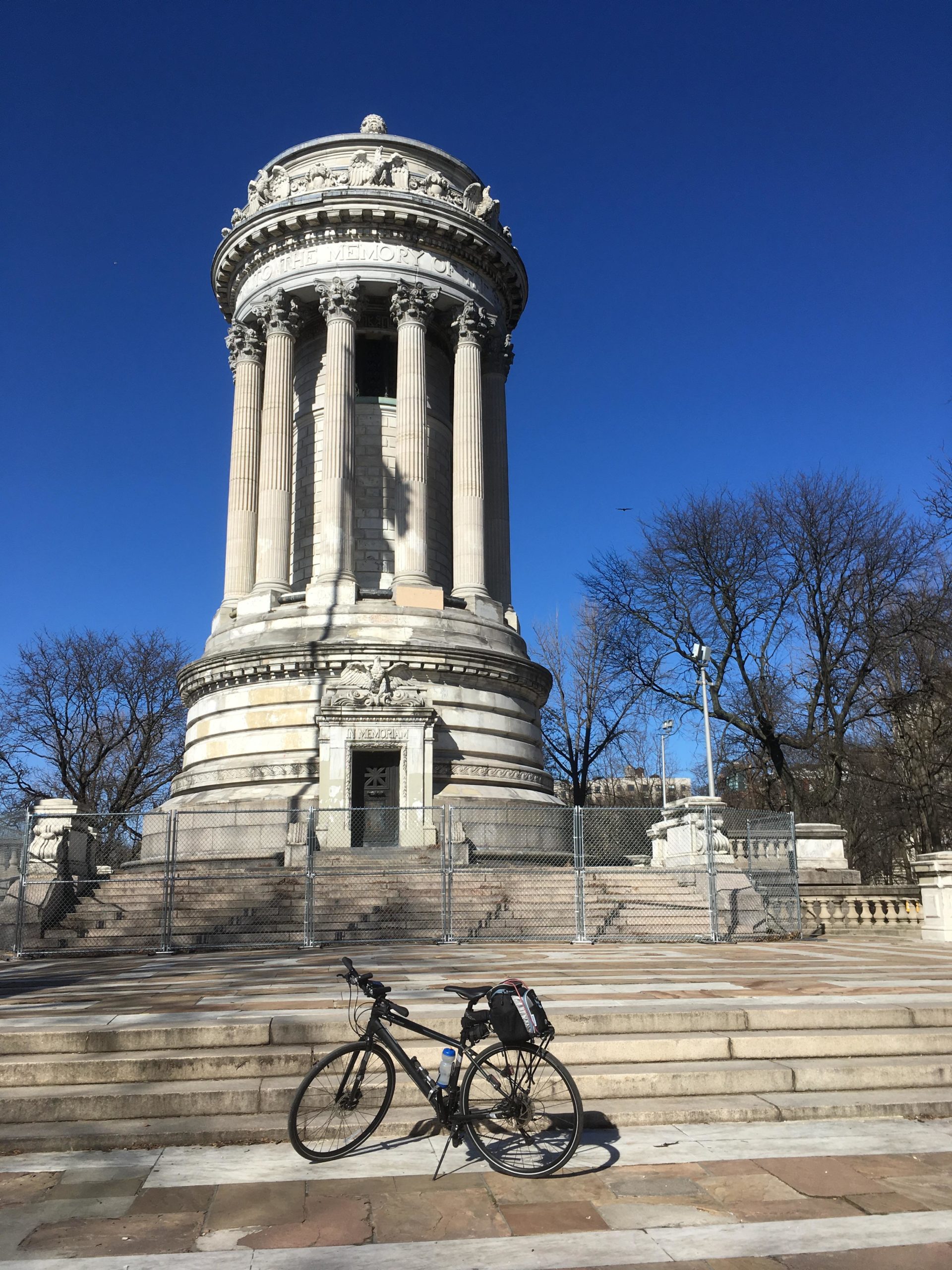 A black bicycle with a water bottle and bag is positioned on the stairs in front of a large, classical monument with columns, under a clear blue sky. The monument features detailed sculptures and inscriptions, surrounded by a temporary fence. Leafless trees can be seen in the background. West Street Greenway mountain bike trail.