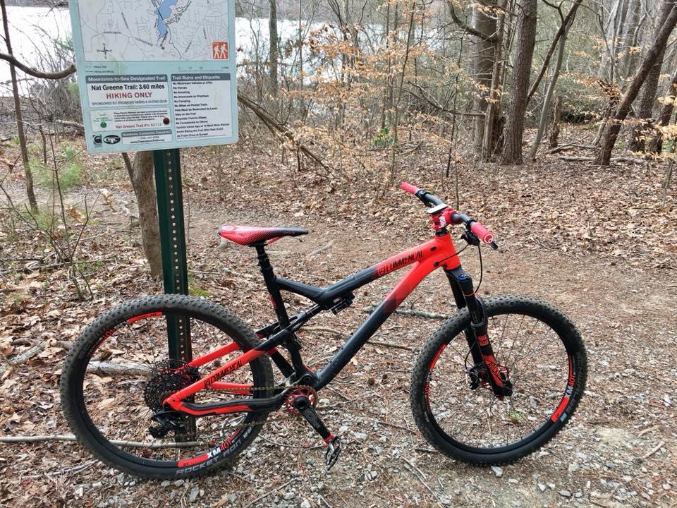 A mountain bike in black and red colors is parked beside a sign for the Nat Greene Trail, indicating a 3.60-mile hiking route. The background features a wooded area with sparse leaves, and a body of water is visible in the distance. Reedy Fork mountain bike trail.