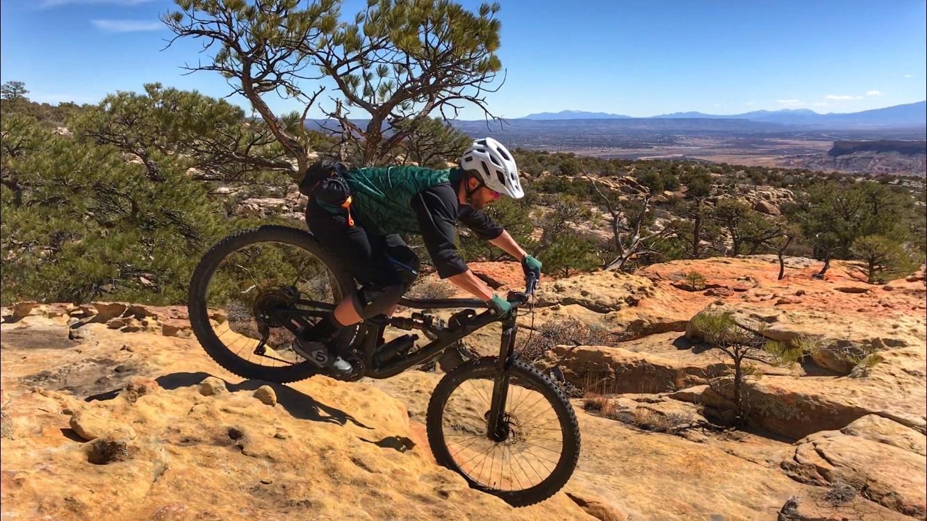 A young person riding a mountain bike over rocky terrain, surrounded by sparse trees and a scenic landscape in the background, under a clear blue sky. Red Mesa mountain bike trail.