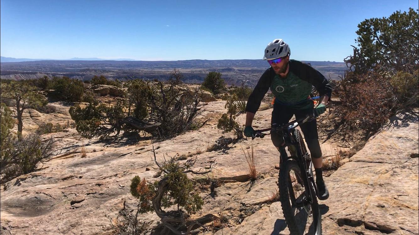 A mountain biker navigating a rocky trail with expansive desert scenery in the background. The sun is shining, and the sky is clear, highlighting the natural landscape. The biker is wearing a helmet and sunglasses, dressed in a sports outfit suitable for cycling. Red Mesa mountain bike trail.