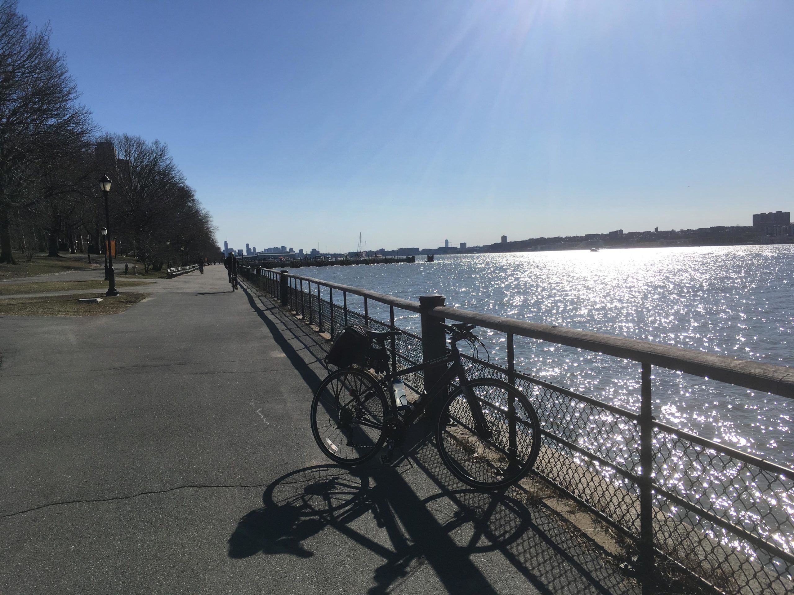 A sunny day along a waterfront path, featuring a bike leaning against a railing. The path runs parallel to a shimmering body of water, with silhouetted trees on the left and a city skyline visible in the distance. West Street Greenway mountain bike trail.