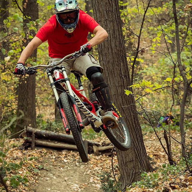 A mountain biker in a red shirt and helmet jumps off a dirt ramp in a forested area, surrounded by trees and autumn foliage. The biker is mid-air, showing dynamic action, with bike tires lifted off the ground and an intense focus on the trail ahead. Bike Park Josutnjak mountain bike trail.