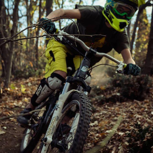 A young person in protective gear rides a mountain bike on a trail surrounded by autumn foliage. The rider, wearing bright yellow shorts and a helmet, is navigating a bend, with a branch partially visible in the foreground. Dust is kicked up behind the bike, indicating speed and movement through a forested area. Bike Park Josutnjak mountain bike trail.