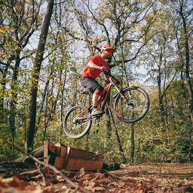 A person in a red shirt and helmet performs a jump on a mountain bike over a wooden ramp in a forested area during autumn. The surrounding trees are displaying fall foliage, with leaves scattered on the ground. Bike Park Josutnjak mountain bike trail.