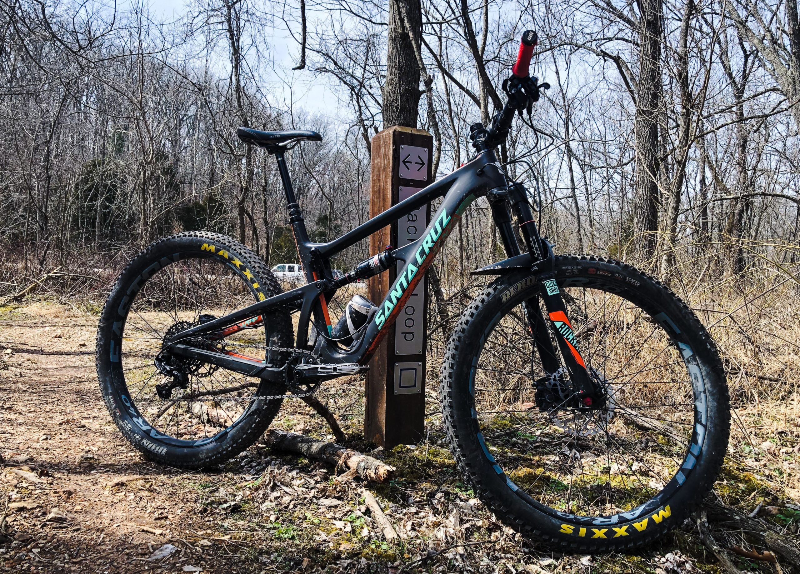 A mountain bike leaning against a trail sign in a wooded area, with bare trees and a gravel path visible in the background. The bike features a black frame with colorful accents and large, knobby tires. Back 40 mountain bike trail.